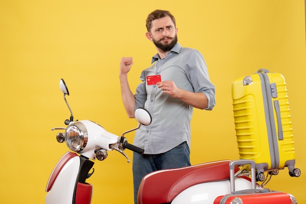 Concetto di viaggio con giovane ambizioso in piedi dietro la motocicletta con le valigie su di esso e che tiene la carta di credito su giallo