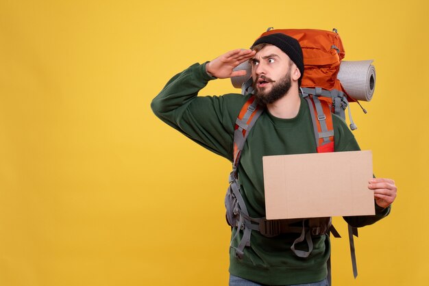 Travel concept with concentrated young guy with packpack and holding free space for writing looking up on yellow 