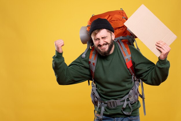 Travel concept with happy emotional young man with packpack and holding a sheet without writing on it on yellow 