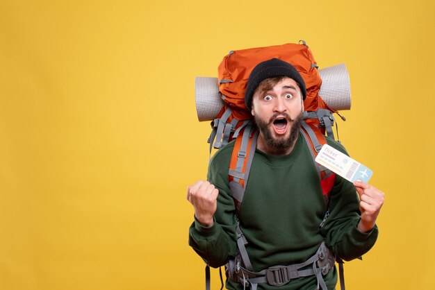 Travel concept with nervous emotional young guy with packpack and holding ticket on yellow 