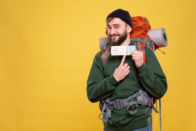 Концепция путешествия с улыбающимся молодым парнем с пакетом и указывающим билетом на желтом