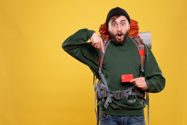 Travel concept with surprised young guy with packpack and holding bank card pointing down on yellow 