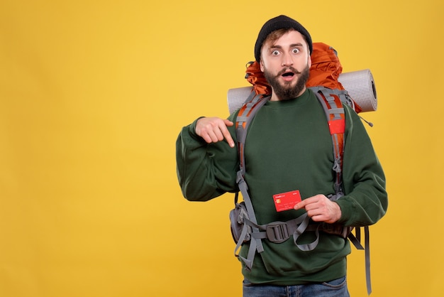 Travel concept with wondering young guy with packpack and pointing bank card on yellow