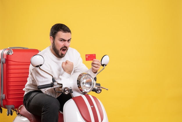 Foto gratuita concetto di viaggio con un giovane ambizioso uomo in viaggio emotivo seduto su una motocicletta con la valigia sopra che tiene una carta bancaria su giallo