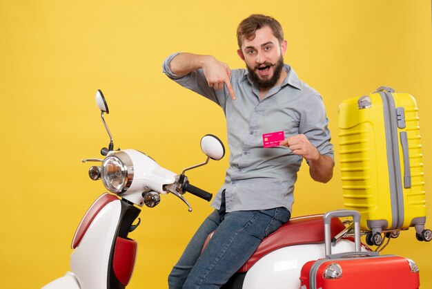 Travel concept with young confident bearded man sitting on motocycle pointing bank card on it on yellow 