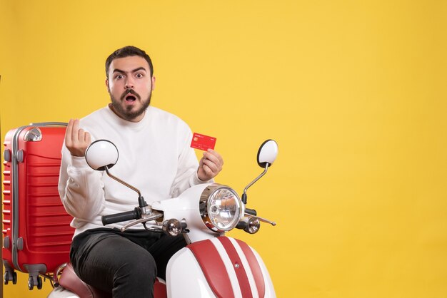 Travel concept with young confused travelling man sitting on motorcycle with suitcase on it holding bank card on yellow