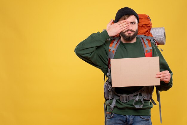 Travel concept with young guy with packpack and pointing free space for writing put his hand on one of his eye on yellow 