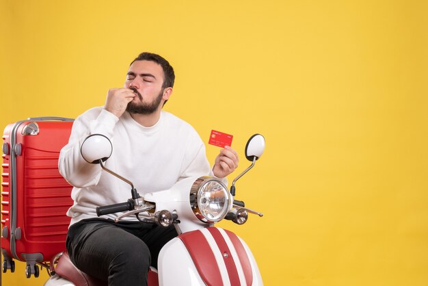 Travel concept with young happy travelling man sitting on motorcycle with suitcase on it holding bank card making perfect gesture on yellow