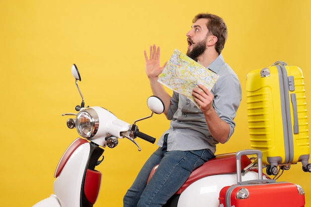 Concetto di viaggio con il giovane uomo barbuto nervoso che si siede sulla motocicletta e che mostra la mappa su di esso su giallo