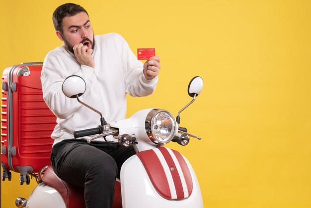 Travel concept with young surprised travelling man sitting on motorcycle with suitcase on it holding bank card on yellow