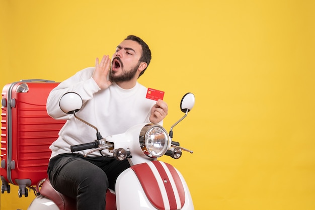 Travel concept with young travelling man sitting on motorcycle with suitcase on it holding bank card calling someone on yellow