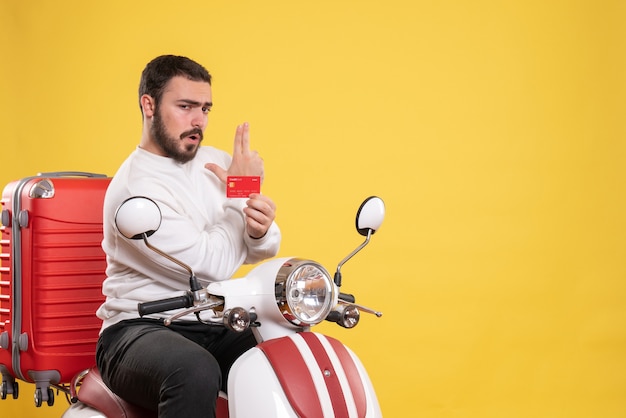 Travel concept with young travelling man sitting on motorcycle with suitcase on it holding bank card making gun gesture on yellow