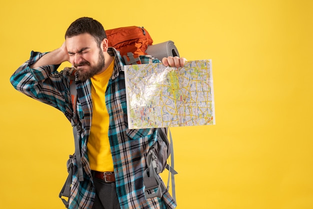 Foto gratuita concetto di viaggio con un giovane ragazzo in viaggio problematico con uno zaino che tiene in mano una mappa che soffre di dolore al collo su giallo