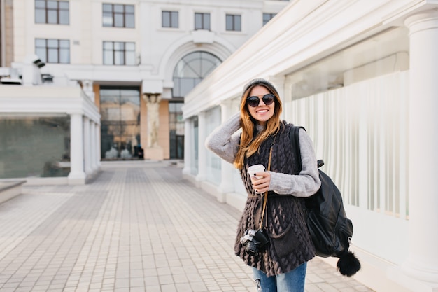 Travel happy time in modern city centre of yoyful pretty young woman in sunglasses, warm winter woollen sweater, knitted hat. Travelling with backpack, coffee to go, camera. Place for text.