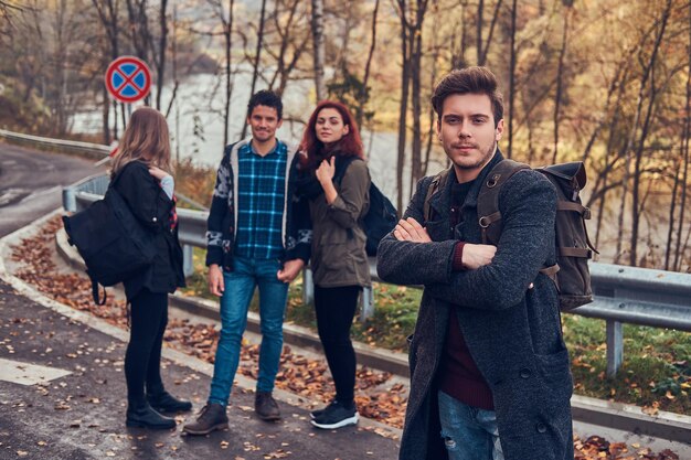 Travel, hitchhiking, adventure concept. Group of young hikers standing on the road sidelines at beautiful autumn forest.