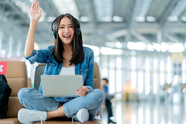 Travel vacation conceptpov asian female woman wear headphone hand use laptop hand wave greeting to camera with cheerful and happinessasian female sit wating at wating area in airport terminal