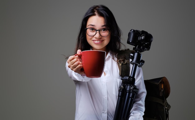 Traveler female holds digital photo camera and a red coffee cup over grey background.