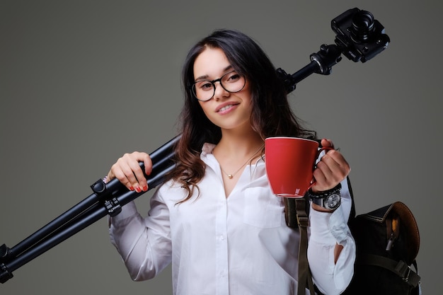 Женщина-путешественница держит цифровой фотоаппарат и красную кофейную чашку на сером фоне.
