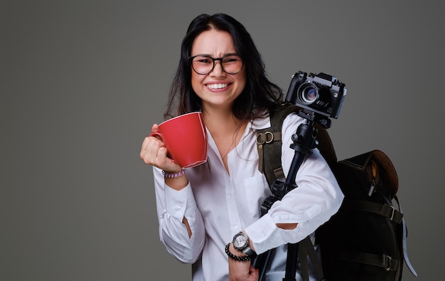 여행자 여성은 회색 배경 위에 디지털 사진 카메라와 빨간 커피 컵을 들고 있습니다.
