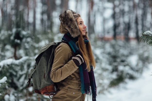 Traveler girl in warm winter jacket with fur hood and big rucksack walking in forest 
