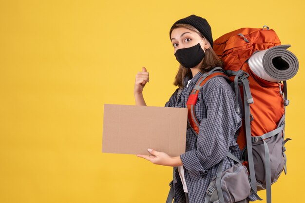 Девушка-путешественница с черной маской и рюкзаком держит картон, указывая на спину на желтом