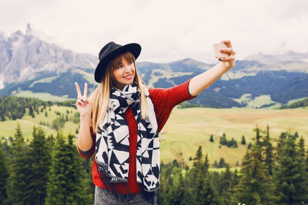 Traveler stylish young woman uses smartphone for taking self photo on mountains