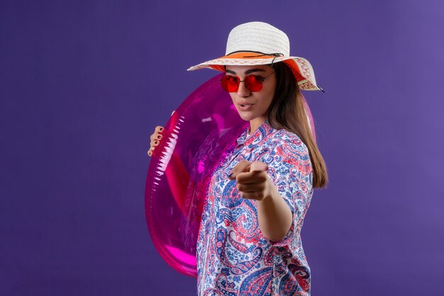 traveler woman wearing summer hat and red sunglasses holding inflatable ring pointing with index finger with serious and confident facial expression standing on purple