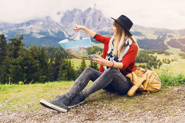 バックパックと帽子の草の上に座って、地図上で正しい方向を検索する旅行者の若い女性