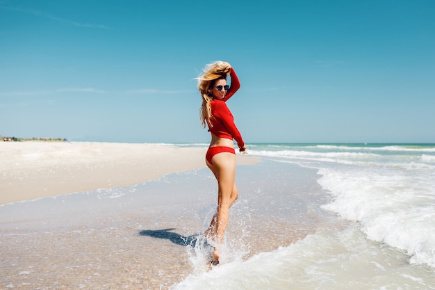 Traveling blond woman jumping with happy expression , turns around in water. Wearing red bikini. Full length. Summer vacation.