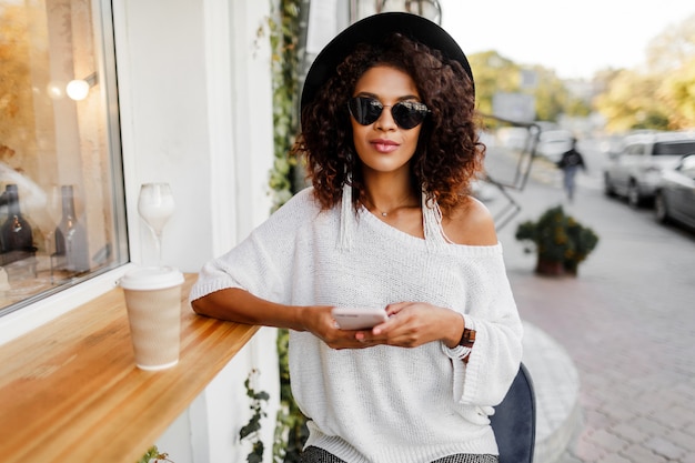 Traveling mix race woman in stylish casual outfit relaxing outdoor in city cafe, drinking coffee and chatting by mobile phone.