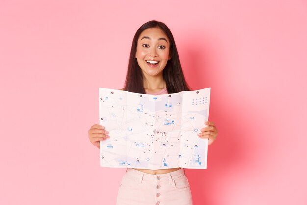 Travelling, lifestyle and tourism concept. Cheerful, attractive asian girl tourist explore new city, visiting museums, showing map of city with sightseeings and smiling upbeat, pink background.