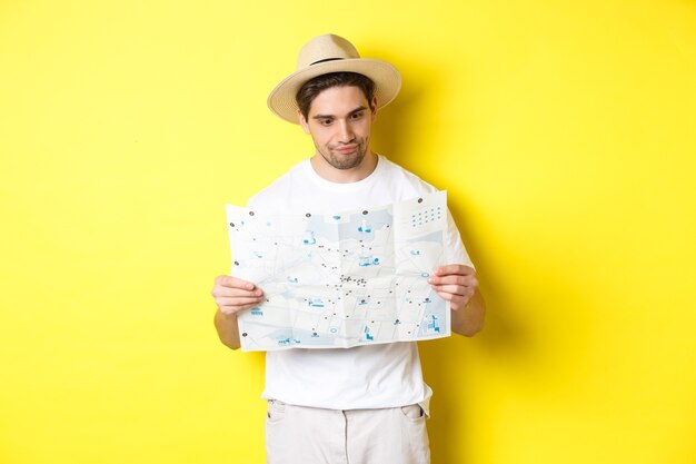 Travelling, vacation and tourism concept. Male tourist looking confused at map, looking for right direction, standing over yellow background.