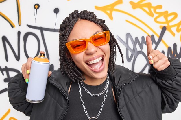 Trendy Afro American teenage girl smiles broadly makes yo gesture poses in urban place uses aerosol spray for drawing graffiti wears sunglasses and jacket belongs to hooligan gang