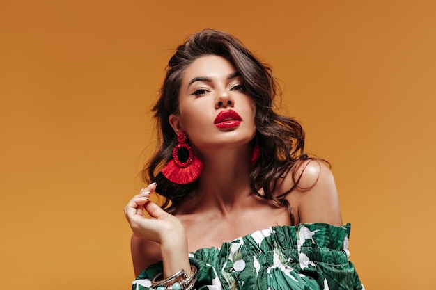 Trendy attractive woman with wavy hairstyle with modern cool earrings and big red lips in green sundress looking into camera