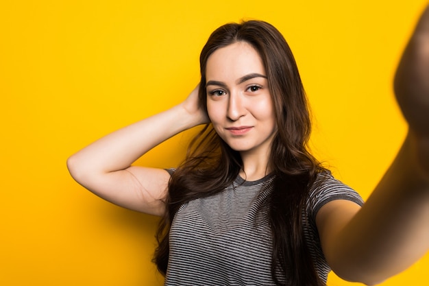 Trendy beautiful girl taking selfie with mobile phone against a yellow wall.