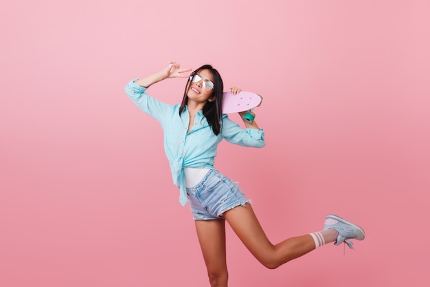 Trendy black-haired woman with suntan dancing with pink longboard and laughing. Sporty asian girl in blue shirt and sunglasses standing on one leg with peace sign.