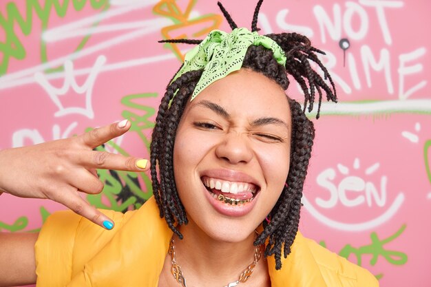 Trendy cheerful hipster girl makes yo gesture shows golden teeth and tongue winks eye enjoys cool modern music stands near graffiti wall enjoys her freedom