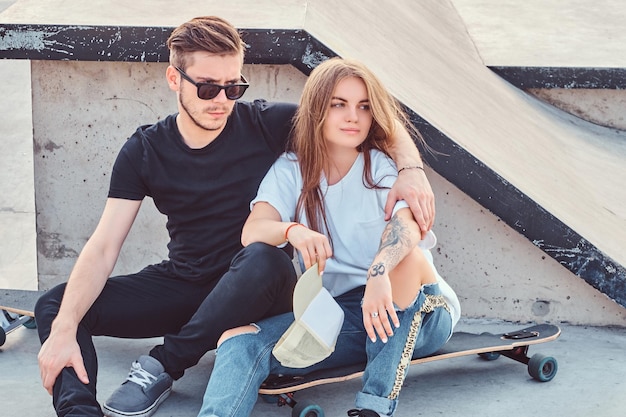 Модно одетая пара молодых фигуристов обнимается, сидя на скейтбордах в скейтпарке в солнечный день.