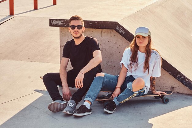 Trendy dressed couple of young skaters sitting on skateboards at skatepark on sunny day.