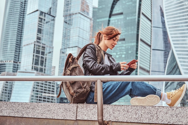 Trendy dressed redhead student girl with tattoos on her face using a smartphone in front of skyscrapers in Moskow city at cloudy morning.