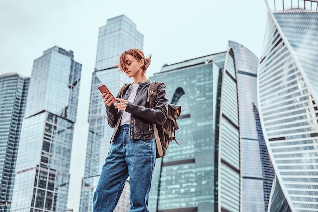 Free photo trendy dressed redhead student girl with tattoos on her face using a smartphone in front of skyscrapers in moskow city at cloudy morning.