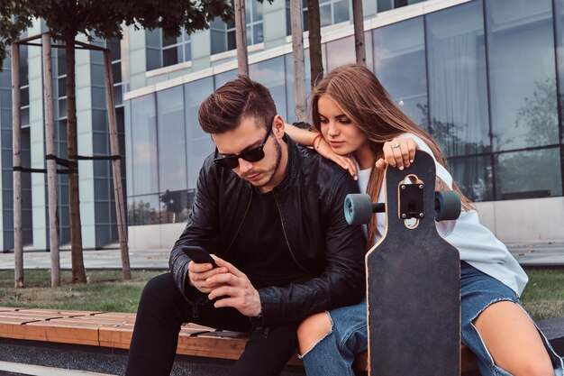 Trendy dressed young couple browsing something on the smartphone while sitting together on bench near skyscraper.