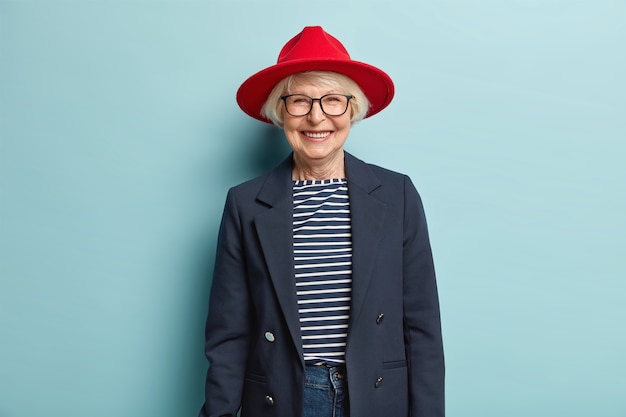 Trendy fashionable elderly woman smiles happily, shows white teeth, has wrinkled skin, dressed in stylish formal clothes, being in good mood, ready for work, enjoys nice day, isolated on blue wall