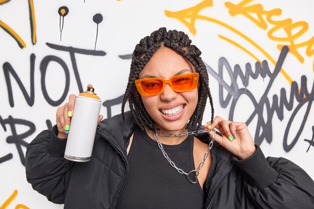 Trendy fashionable teenage girl dressed in black clothes orange sunglasses and metal chain has braid hairstyle poses with aerosol spray makes creative graffiti on street walls