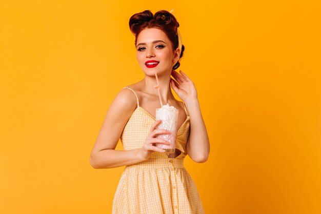 Trendy ginger girl drinking milkshake. Studio shot of fit pinup lady isolated on yellow space.