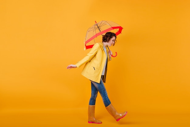 Trendy girl in rubber shoes and yellow jacket looking down while posing with umbrella. Studio shot of curly short-haired woman in jeans walking with parasol.