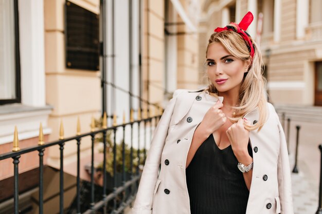 Trendy girl with colorful ribbon in blonde hair playfully posing in cold autumn day standing near store. Outdoor portrait of adorable fair-haired female model wrapped in beige coat .