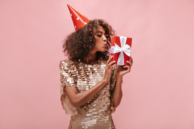 Trendy girl with short brunette hairstyle in beige shiny dress and holiday cap kissing her red gift box on isolated wall..