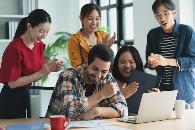 트렌디한 힙스터 아시아의 창의적인 친구는 친구 그룹과 캐주얼한 회의에 앉아 웃고 있으며 노트북 컴퓨터를 디지털로 사용하면서 새로운 성공 프로젝트에서 일하고 있습니다.