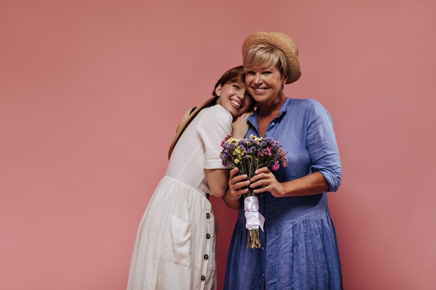 Trendy lady with blonde hairstyle in blue dress and straw hat smiling, holding colorful bouquet and posing with girl in white outfit on pink backdrop. 